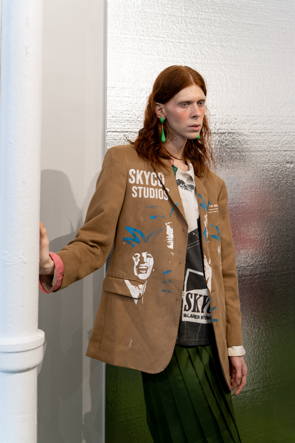 A model stands in a fashion showroom with reflective cardboard behind them. The model is wearing a brown linen jacket with printed patterns and a green dress. The clothing is from the brand Skyco.
