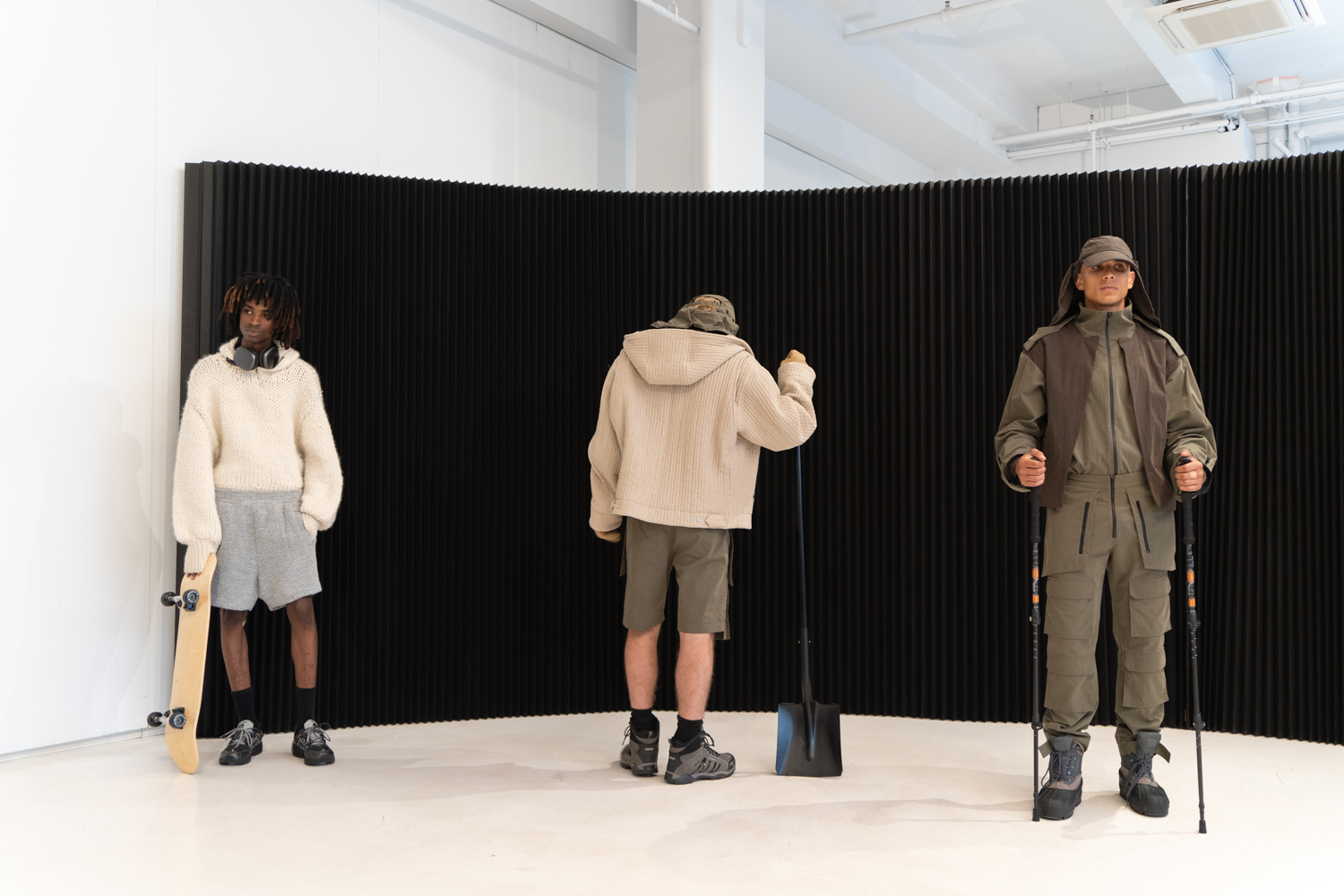 Three models stand in a fashion showroom with a black backdrop and white floors. They are wearing clothing from the brand TARPLEY. The clothing has an outdoor theme. They are holding props including a skateboard, a shovel and trekking poles.