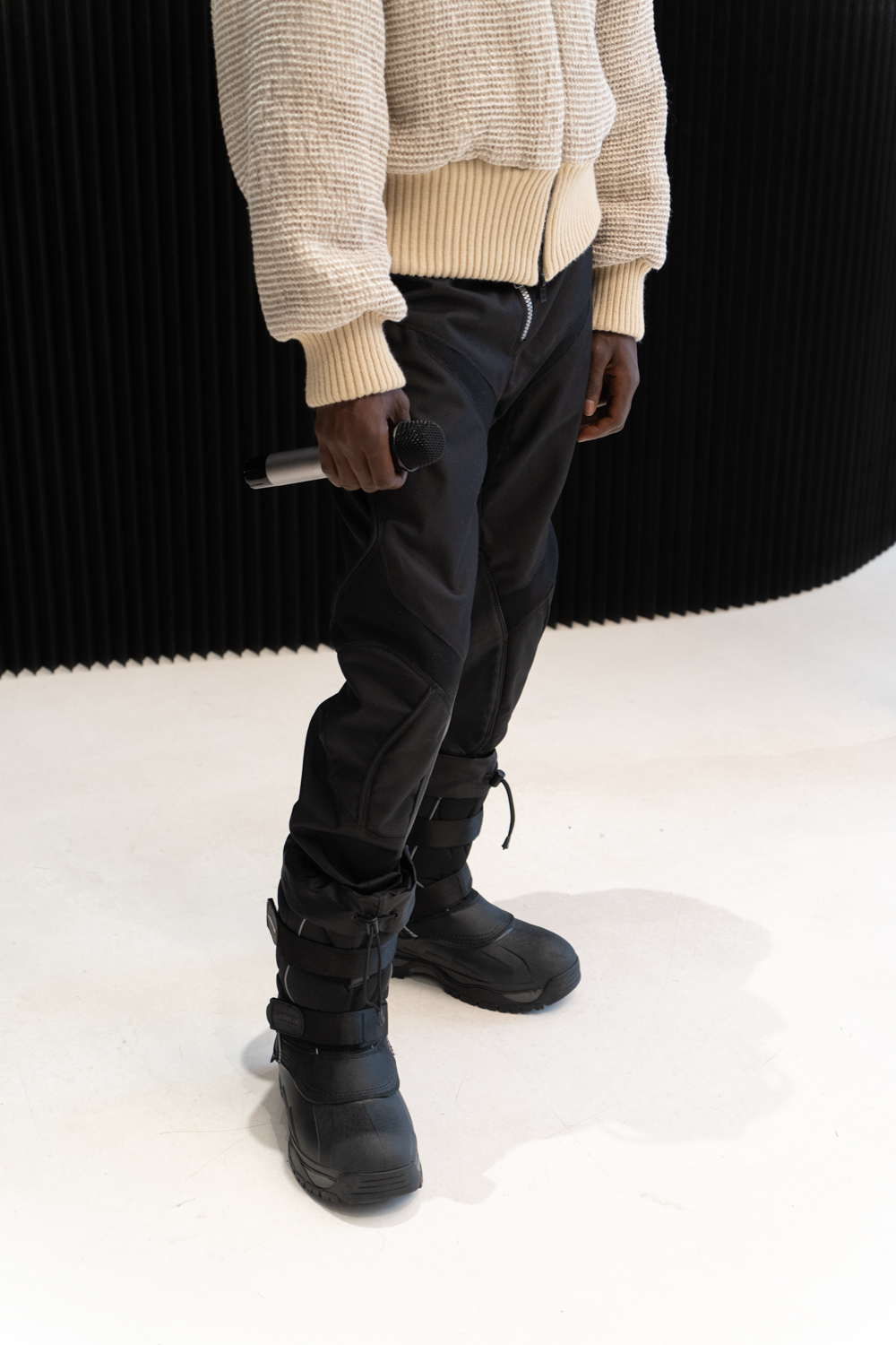 A close-up photo of a model standing in a fashion showroom wearing clothing from the brand TARPLEY. The model is wearing black hiking pants and boots.