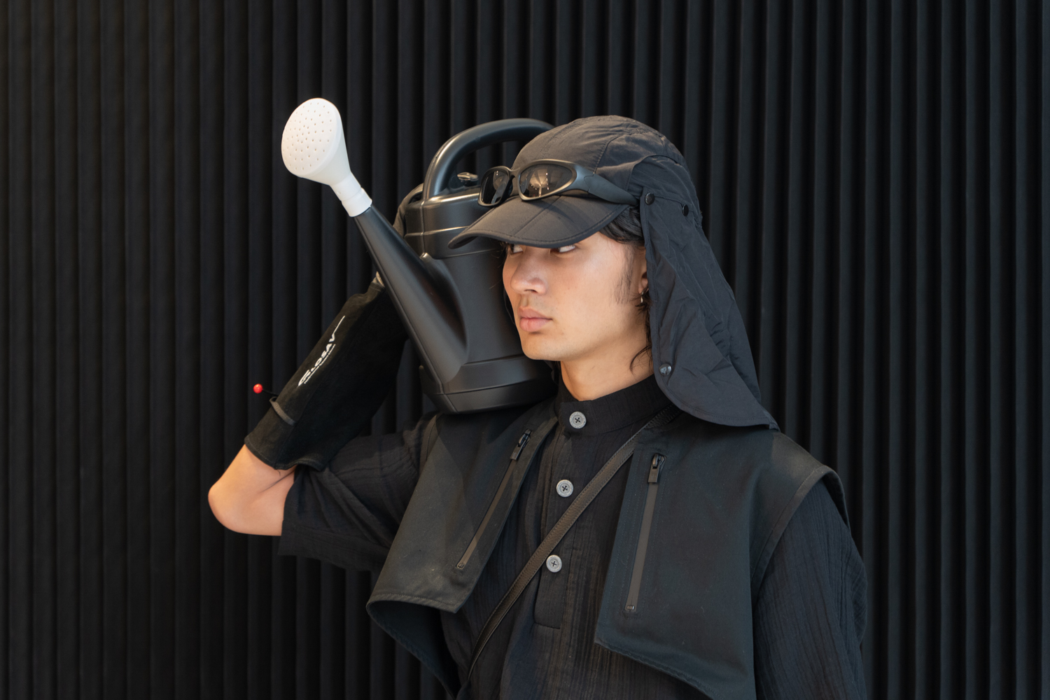 A model stands in a fashion showroom against a black backdrop. The model is wearing clothing from the brand TARPLEY, including a black hat and jacket. The model is holding a watering can.