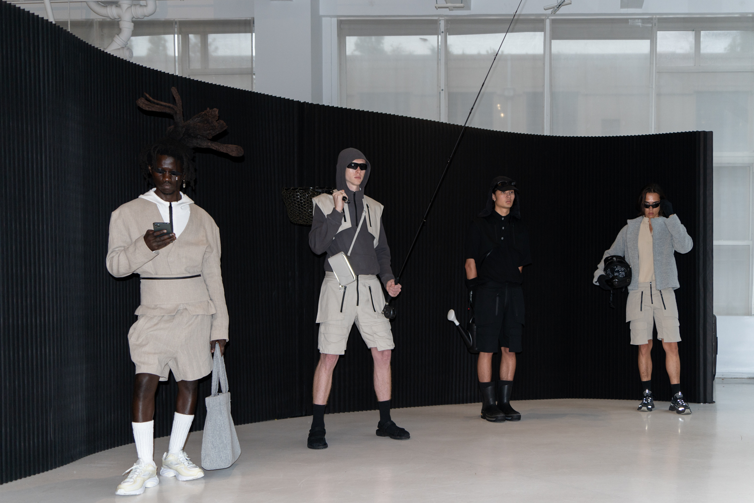Four models stand in a fashion showroom with a black backdrop and white floors. They are wearing outdoor-themed clothing from the brand TARPLEY. They are holding props including a smartphone, a tote bag, a fishing rod, a watering can and a helmet.