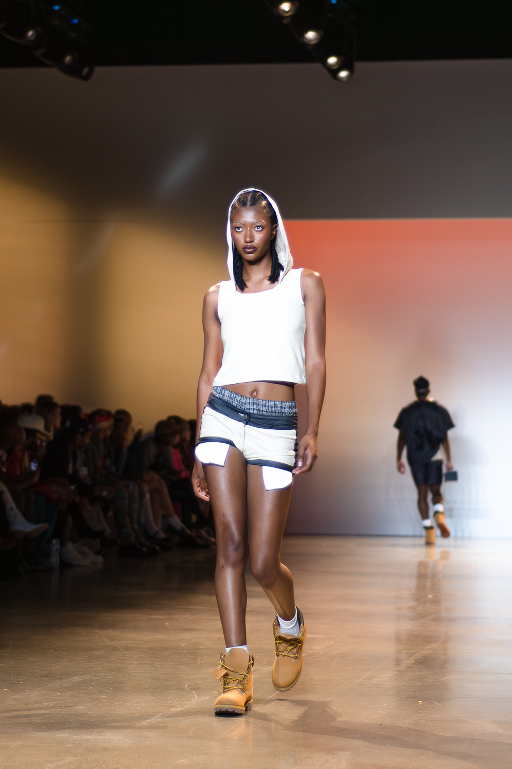 A model wearing a hooded, white tank top with the hood up that stops above the model’s belly button walks on a runway. The model is wearing low-cut short-shorts and brown lace-up boots. The model’s boxers are slightly visible above the shorts.