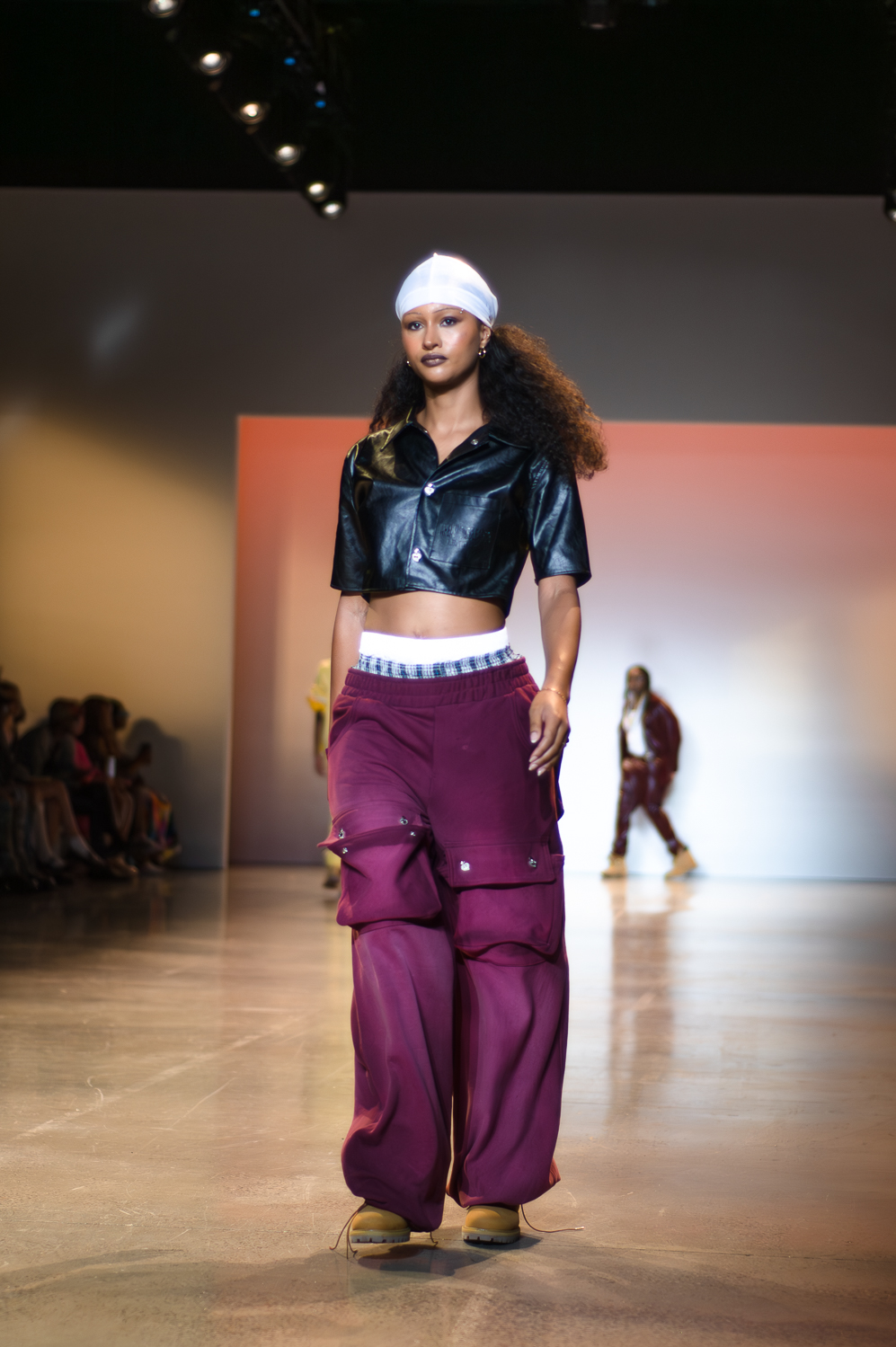 A model wearing a black, seemingly leather, crop top, closed in the front by two buttons, walks on a runway. The model wears a white beanie and baggy, purple pans with big pockets on both legs. The model’s boxers are slightly visible above the pants. The model wears small, gold-colored hoop earrings, and brown lace-up boots.