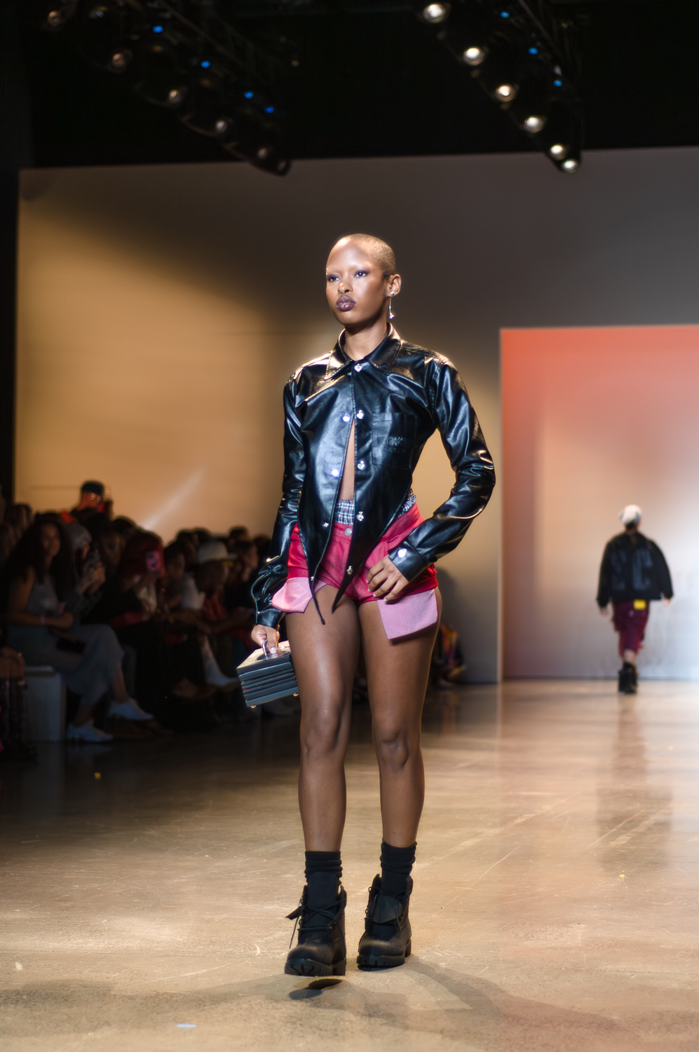 A model wearing a black, leather-like button-up jacket with only the top button closed walks on a runway. The model is wearing short pink shorts, and the model’s boxers are slightly visible above the shorts. The model is wearing black socks and black lace-up boots, and holds a rectangular case by the handle on top.