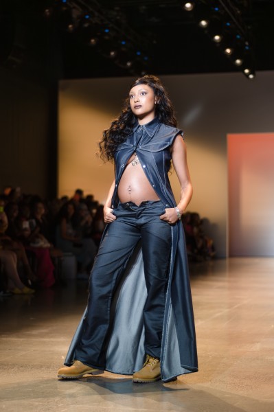A model wearing a floor-length, denim button-up overcoat with only the top two buttons closed and jeans with the fly unbuttoned walks on a runway. The model is pregnant, with the open coat revealing a pregnant stomach. The model wears brown lace-up boots, with thumbs hooked in the pants’ pockets.