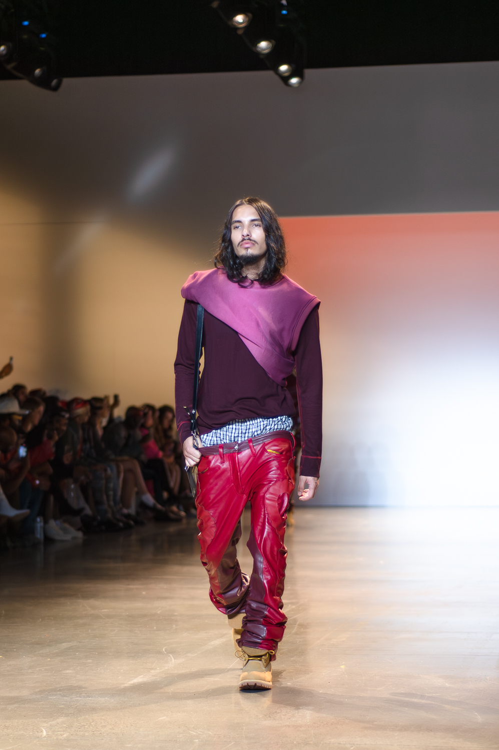 A model wearing a long-sleeve purple shirt and a rolled-up lighter-purple tank top with shoulder pads, which sits diagonally on the model’s chest, walks on a runway. The model is wearing red leather-like pants, and has a bag hanging off one shoulder. The model’s boxers are slightly visible above the pants. The model is wearing brown lace-up boots.
