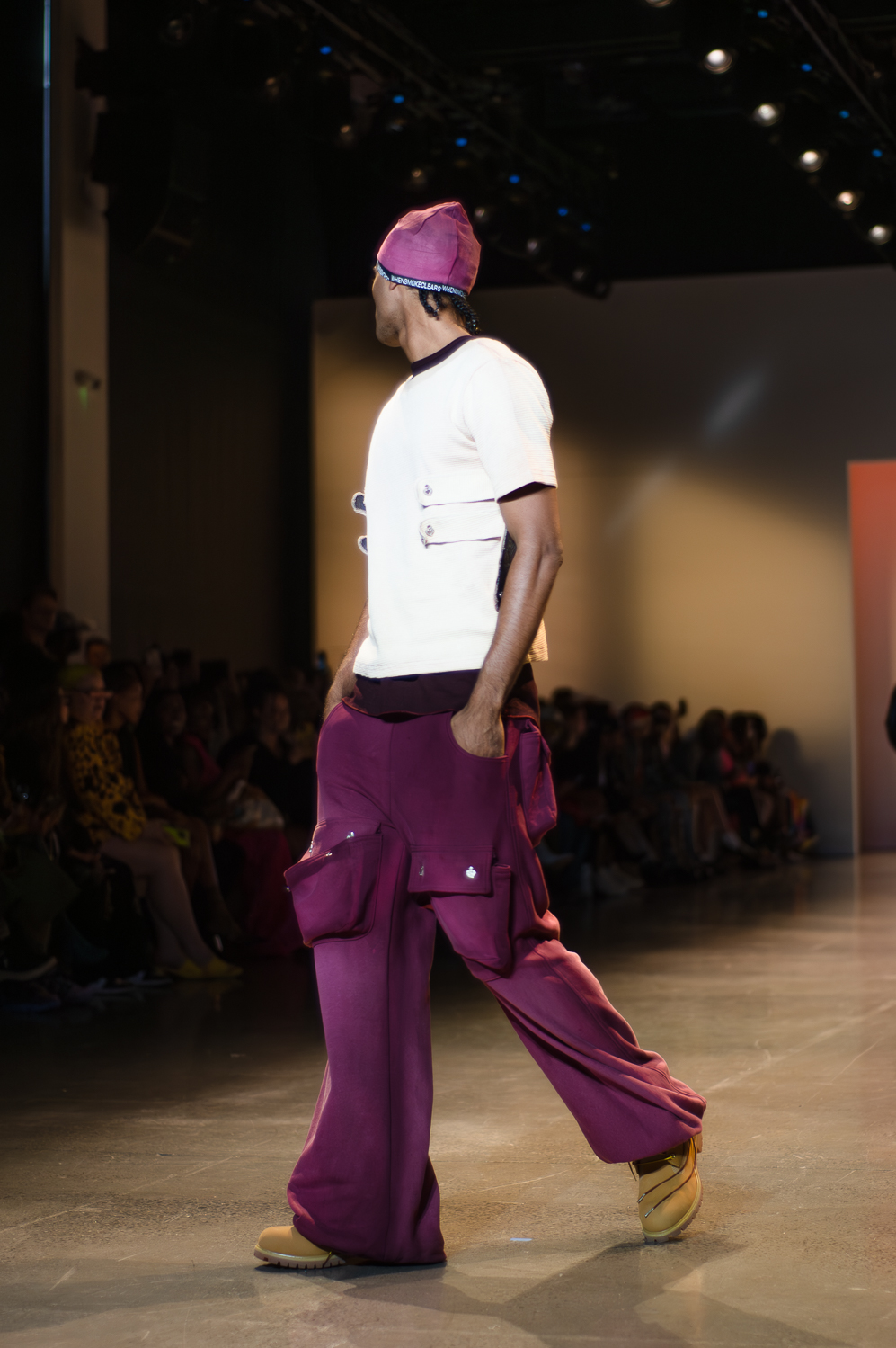 A model wearing a white shirt with two straps on each side of the model’s chest, with a purple shirt underneath, turns to the right on a runway. The model wears baggy purple pants with a large pocket on each leg, and brown lace-up boots. The model wears a pink head covering, and holds both hands in the pants’ pockets.