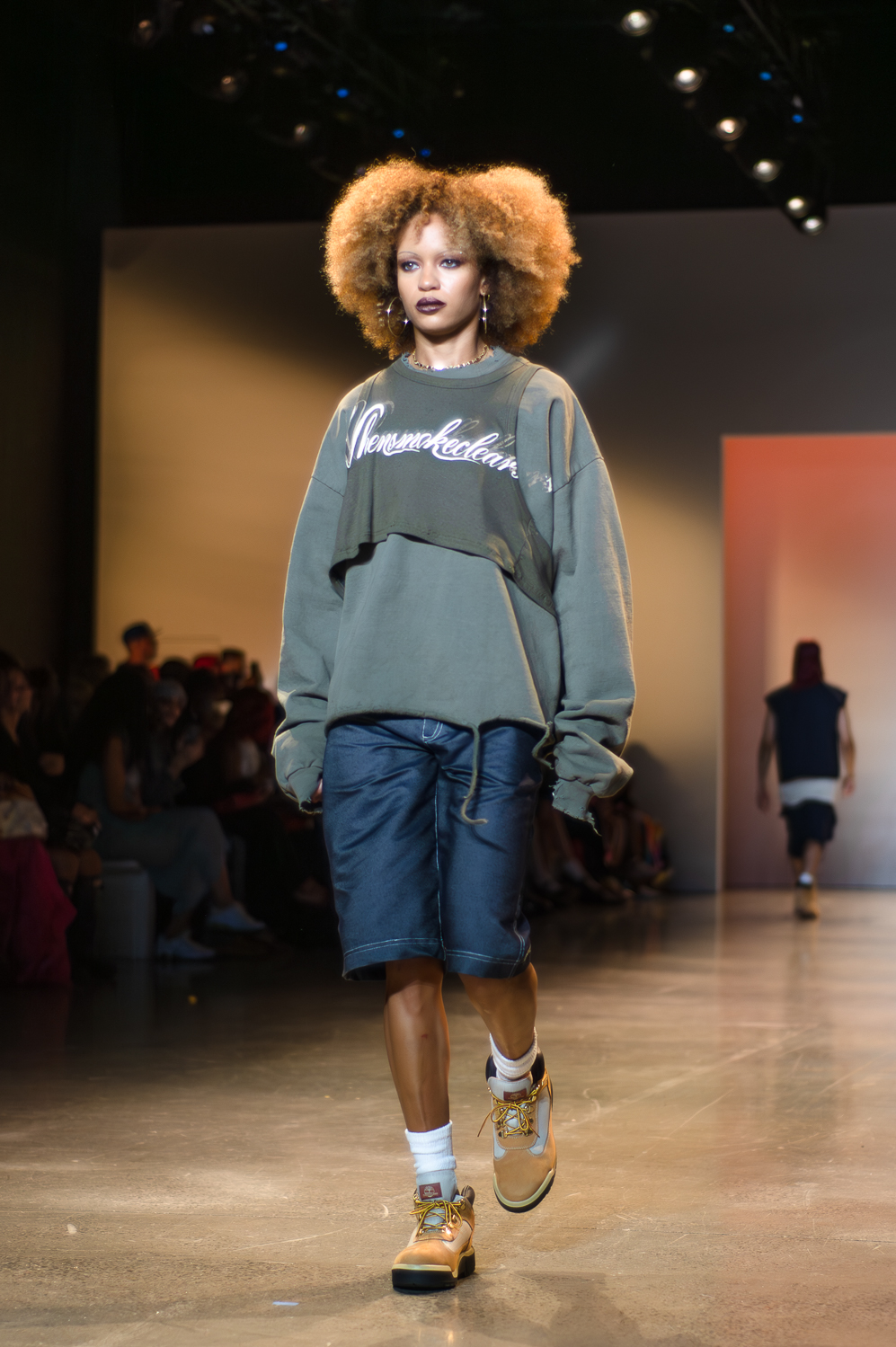 A model wearing a gray sweatshirt with a darker gray tank-top over only her chest with white text on it, and denim capris walks on a runway. The model wears big, gold-colored hoop earrings, a brown almost-choker necklace, and brown-and-gray lace-up boots.