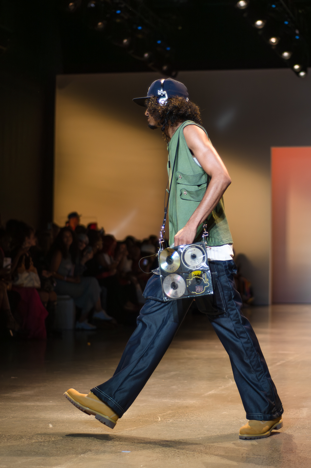 A model wearing a green, flowy tank top, white undershirt, and black pants turns toward the right while walking on a runway. The model wears a blue baseball cap, a headset, and brown lace-up boots. Over the model’s shoulder is a bag with four C.Ds visible.
