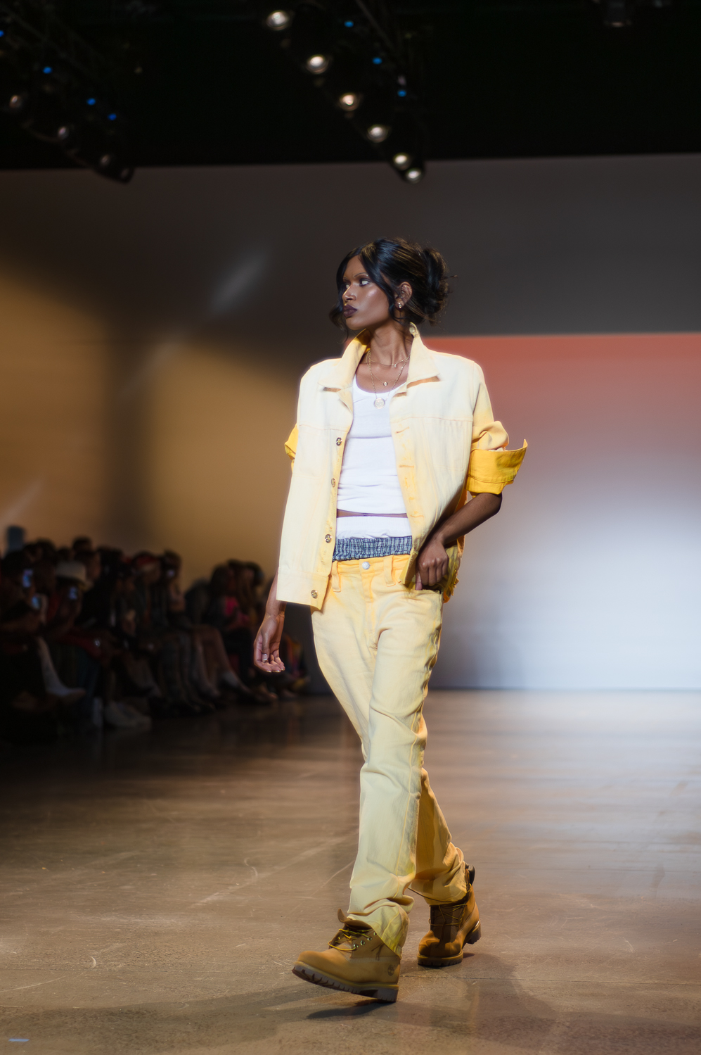 A model wearing a white shirt under a pale yellow, unbuttoned blouse with the sleeves rolled up walks on a runway. The model’s left hand is slightly in the pocket of the matching yellow pants. The model’s boxers are slightly visible above the pants. The model wears a silver necklace with a circle and a shorter silver necklace with a stud-like charm, and brown lace-up boots. The model looks to the right.