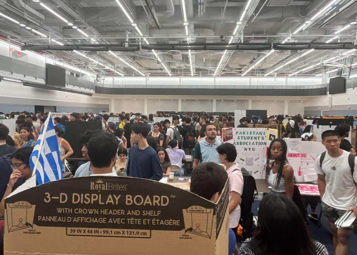 A shot of the Paulson Center gym. Students are standing and looking around. There is a sign for the Pakistani Student’s Association to the right and a Greek flag to the left.