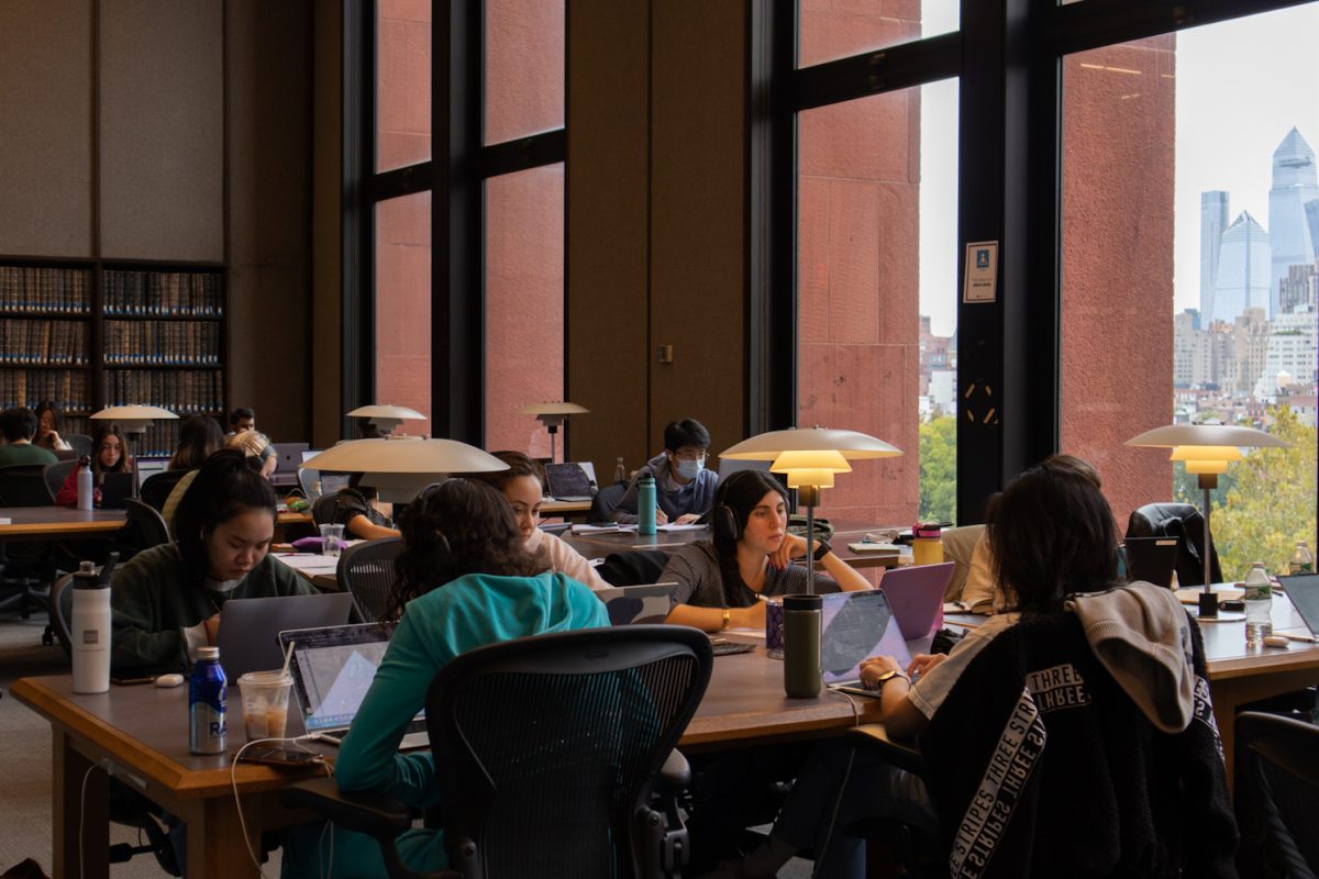 Multiple students sit at long tables working on their computers. To the right is a long window facing Washington Square Park.