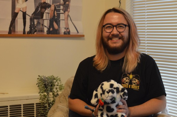 Fino, with shoulder length light pink hair and black glasses, smiles while holding their stuffed toy, a small dalmatian with a rose in its mouth. They are sitting in a corner of their dorm with a poster, plants and window blinds in the background.