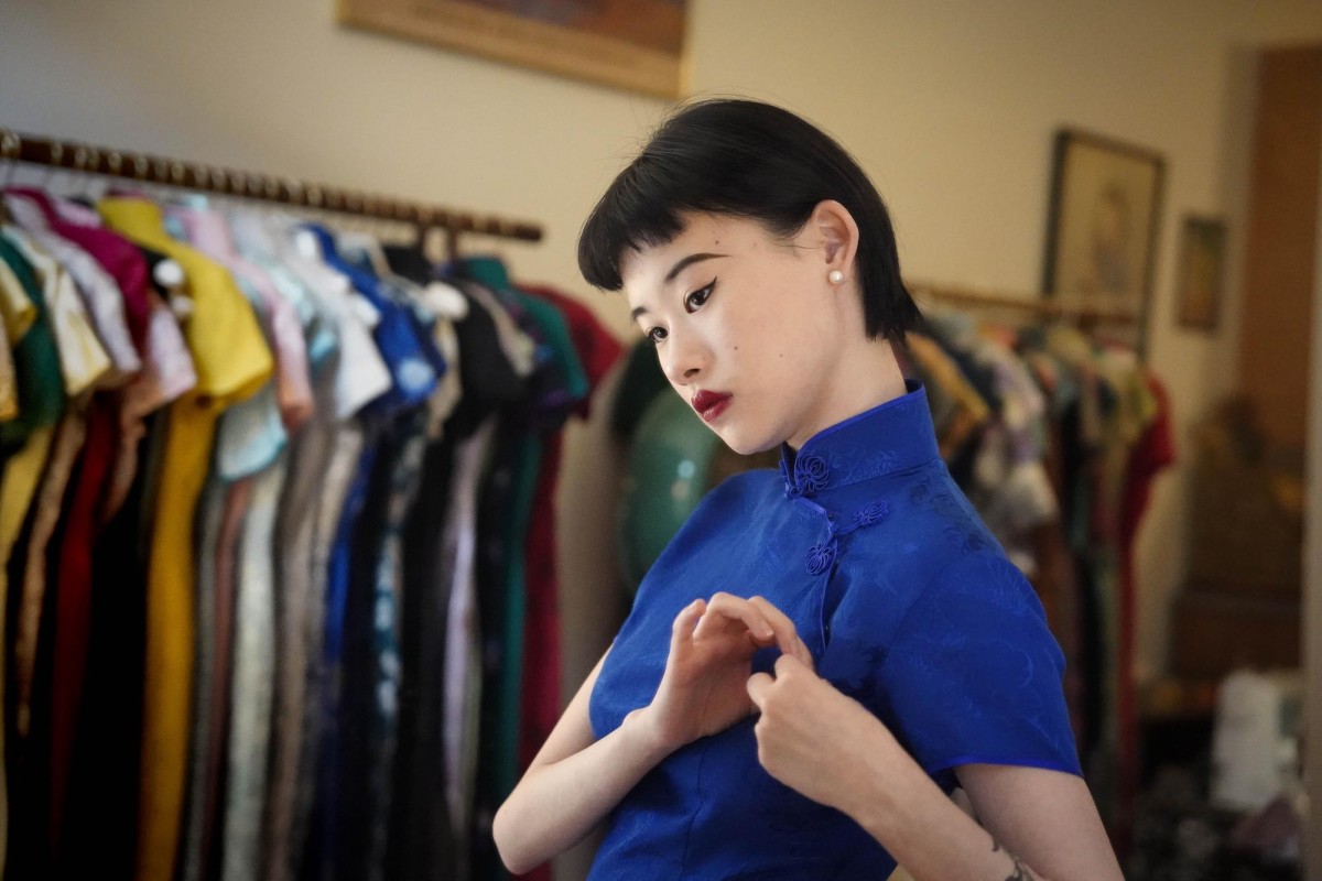 Heather Guo wears a blue cheongsam, showing how to secure it properly. She is standing in front of two clothing racks.