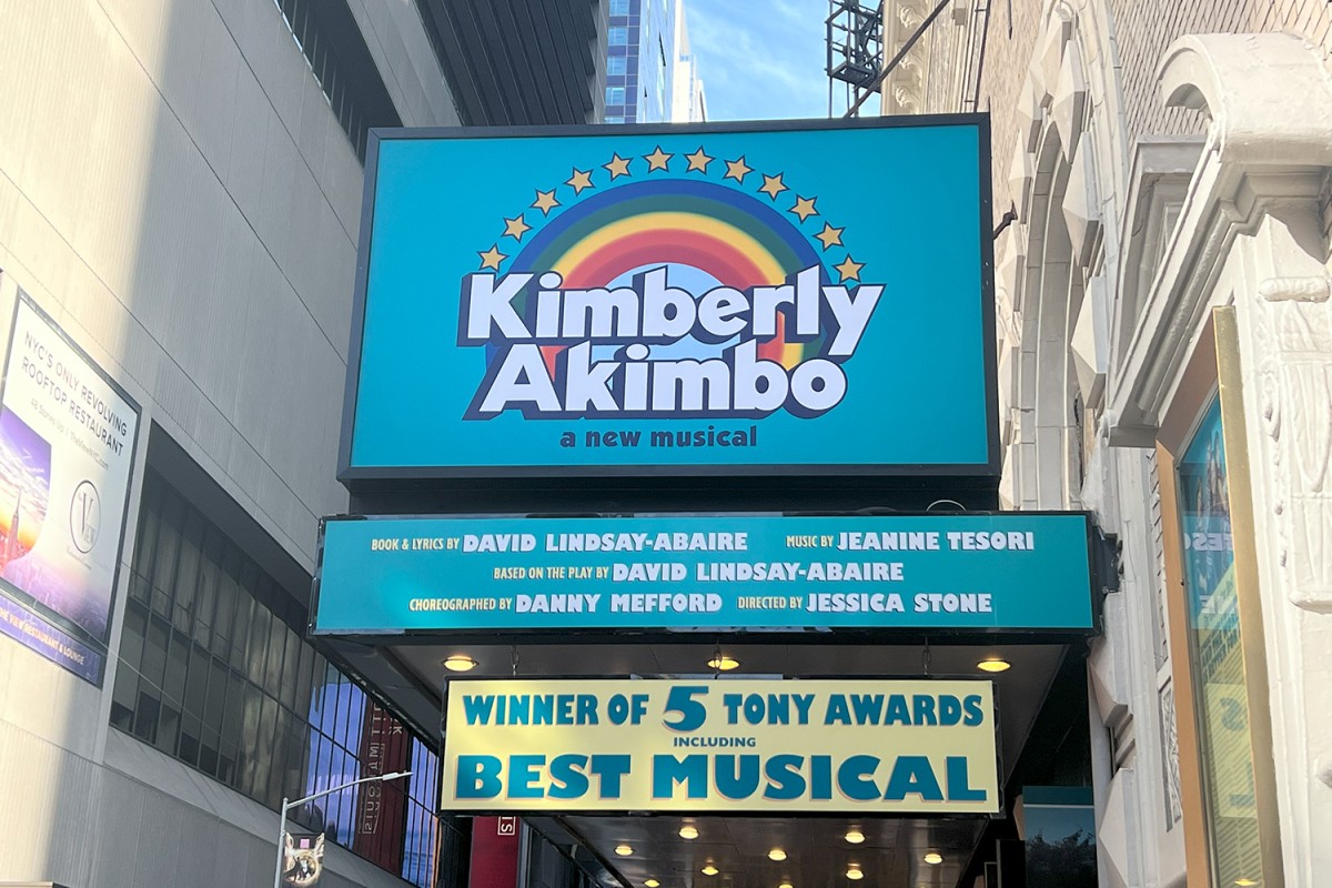 A blue marquee sign with the image of a rainbow outside of the Booth Theatre reading “Kimberly Akimbo.” Below the sign are two smaller signs, the top has a list crediting the creators of the musical and the bottom is a yellow sign that reads “Winner of 5 Tony Awards Including Best Musical”.