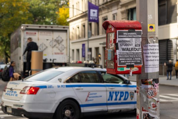 A white paper that “National Student Walkout” posted on the side of a fire box on a light pole. Behind it is a police car and a building with the purple NYU flag.