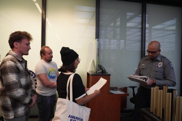 Three people are talking to a campus safety officer. One of them, wearing glasses and a beanie, is holding a piece of paper with text and reading what is written on it.