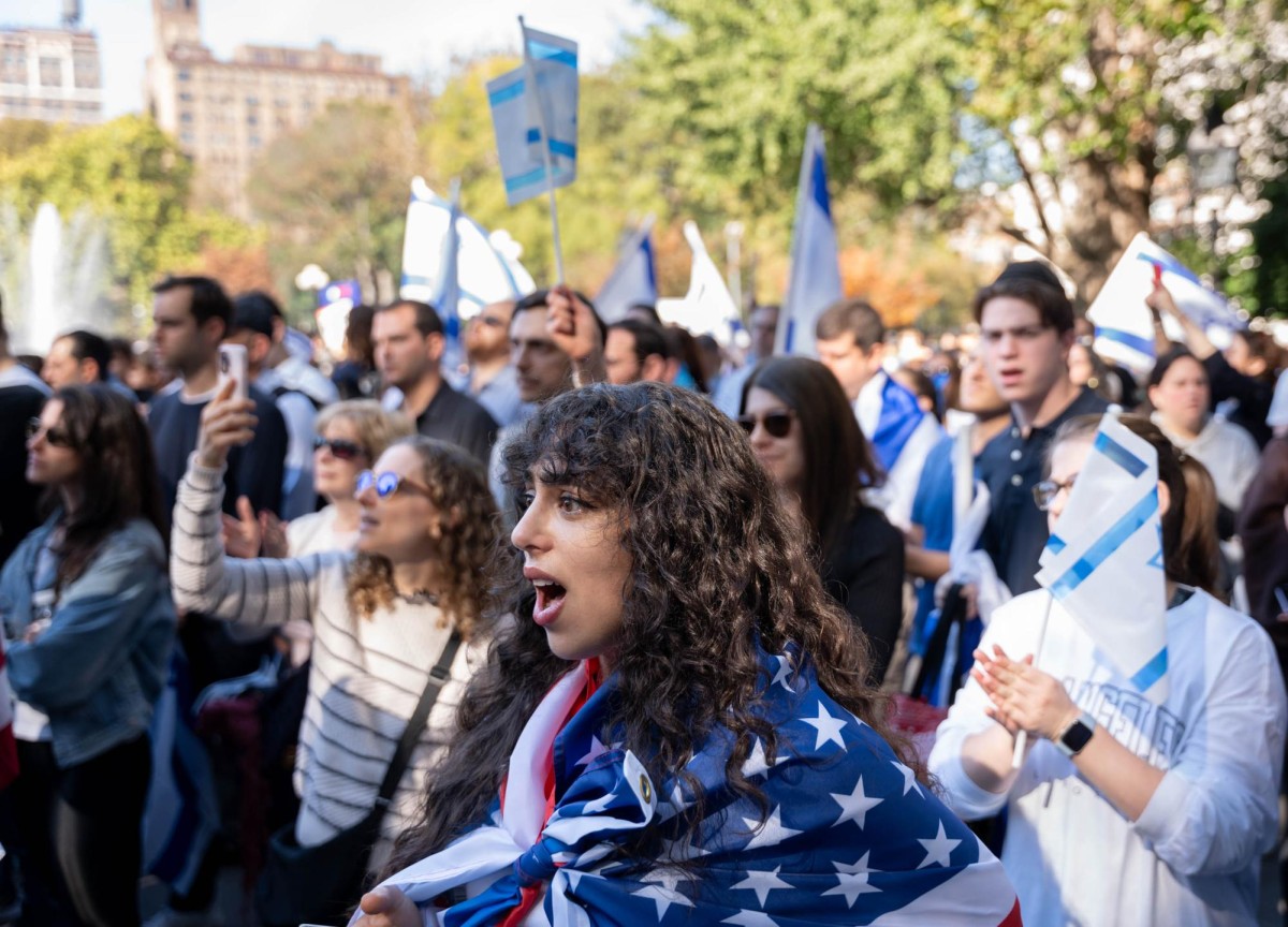 A young person wearing an American flag around stands with a crowd of protestors holding and wearing Israeli flags.