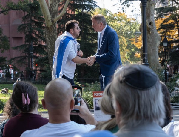 Bill de Blasio shakes hands with a man that is holding a microphone and wearing an Israeli flag around his shoulders.