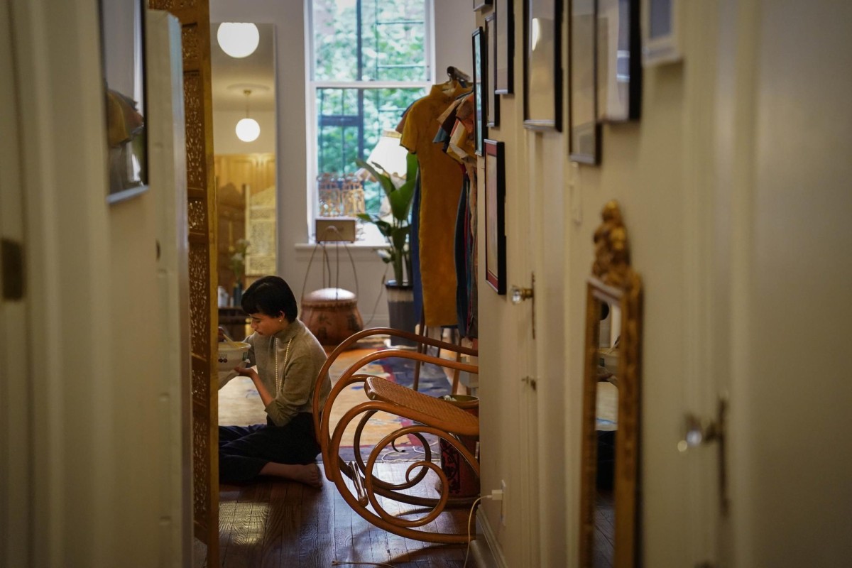 A woman sits on the floor of a room at the end of a hallway, eating a bowl of noodles.