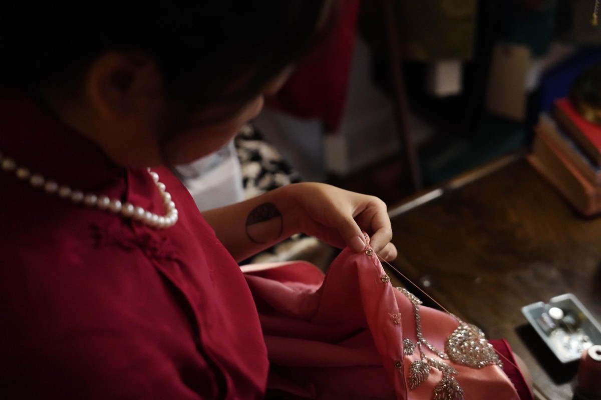 An over-the-shoulder view of a woman in a red dress mending a piece of clothing with a needle and a thread.