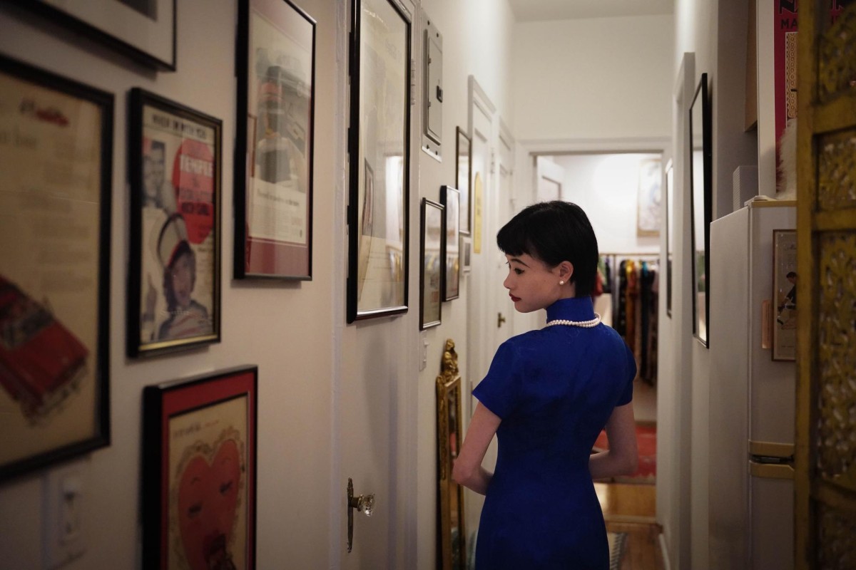 A woman wearing a blue Cheongsam dress glances back in a hallway. The wall by her side is decorated by a collection of vintage magazine covers and posters.