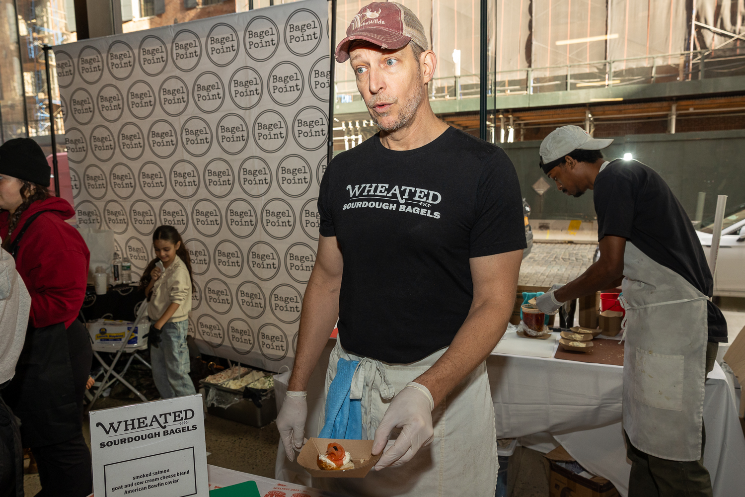 A man in a red baseball hat wearing a black shirt and white apron with white gloves handing out samples.