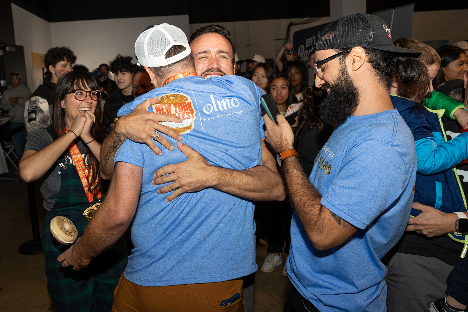 A man in a blue shirt and white hat from Olmo holding a bagel award for “Best of the Fest” and is hugging another man.