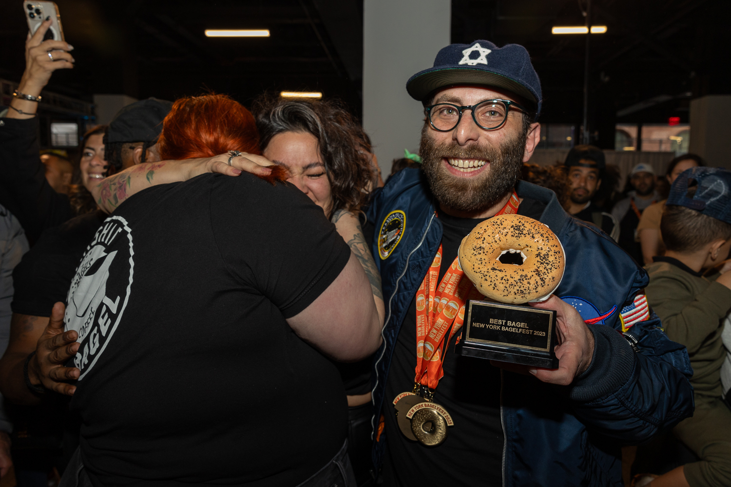 Two women with black shirts hugging. On the right, a man from Starship Bagel, with a black shirt, blue jacket and hat holding a bagel shaped award for “Best Bagel.” He also has three bagel-shaped medals with an orange ribbon around his neck.