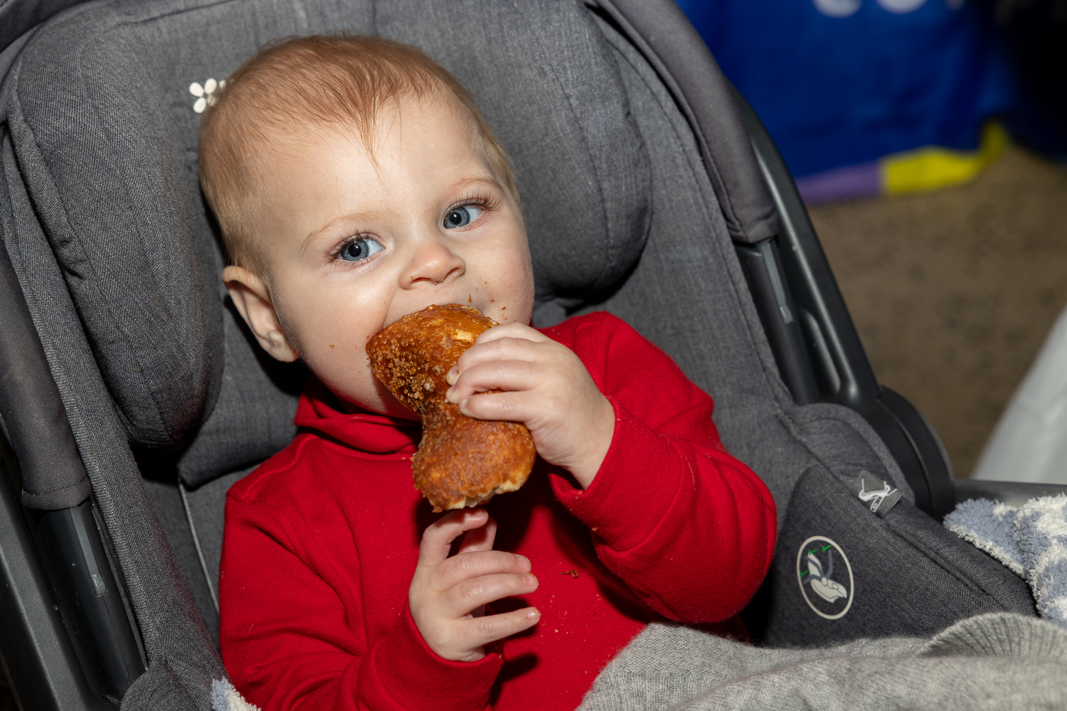 A baby named Madison in a red hoodie eating a bagel in a gray car seat.