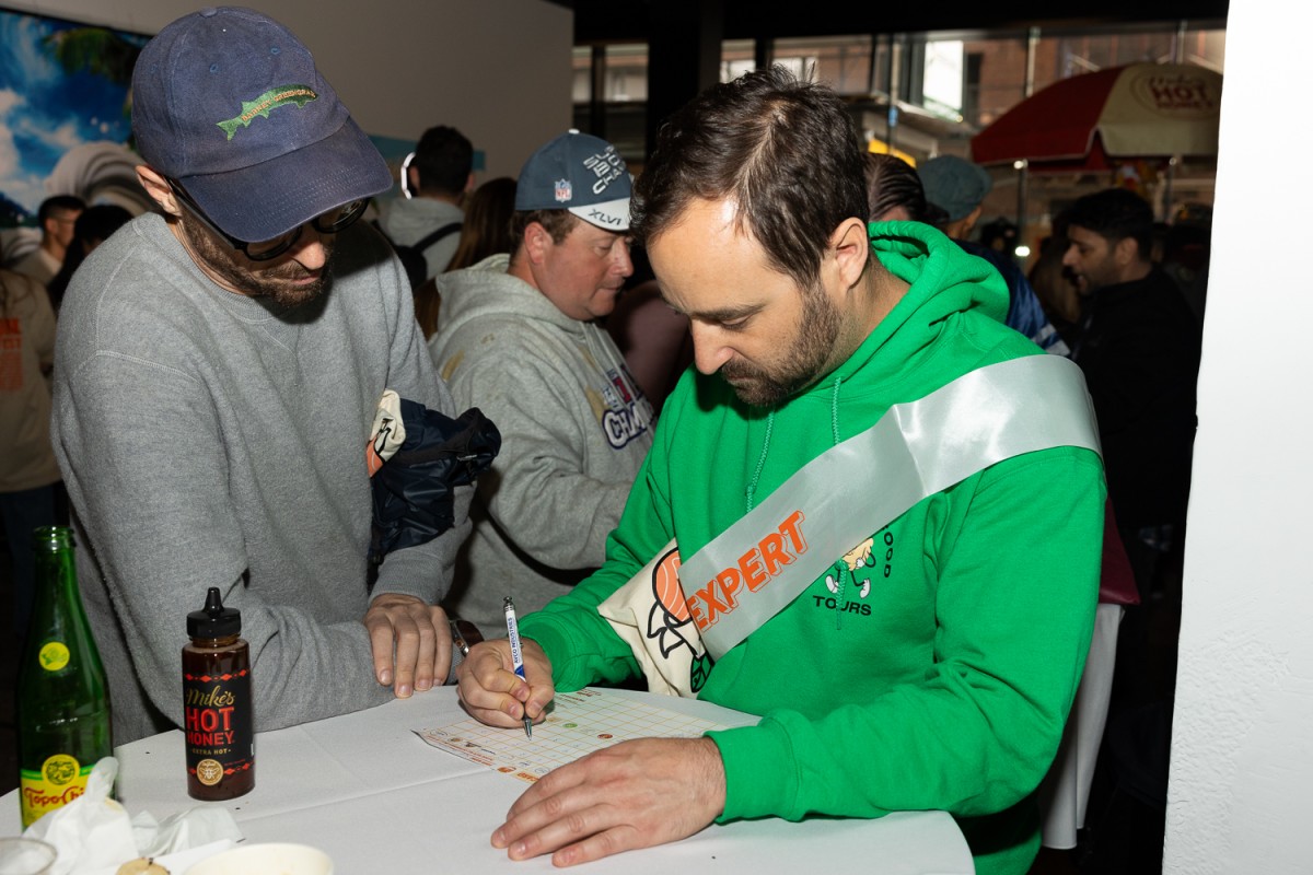Scott Goodfriend, the Chief Eating Officer at Ultimate Food Tours, wearing a green hoodie and “Bagel Expert” sash writing with a pen on a score sheet listing all the vendors.
