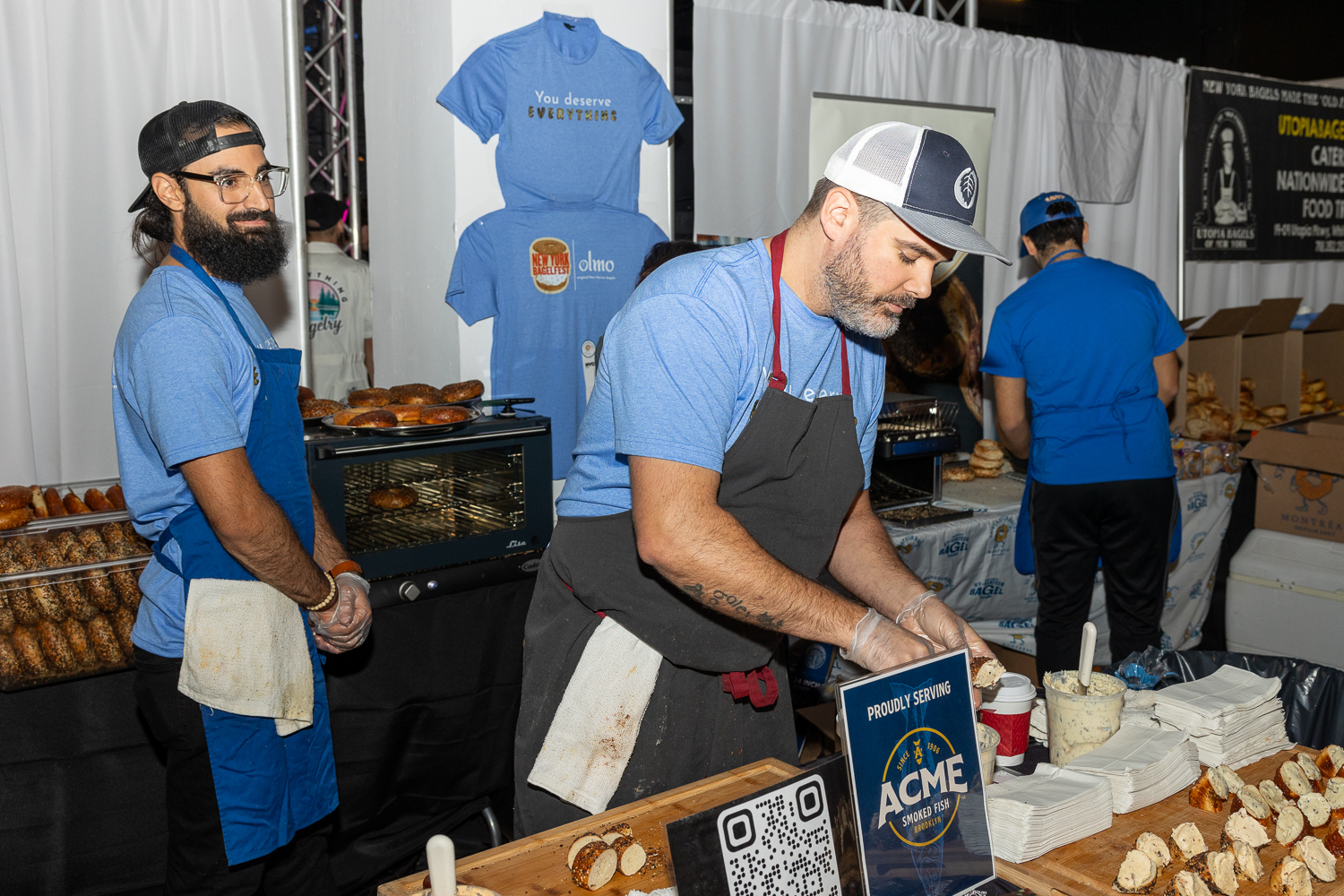A man in a black hat and blue shirt from Olmo standing on the left. Another man in a white-and-gray hat, blue shirt and gray apron, also from Olmo, serving bite-sized everything bagels filled with Yuzu Kosho cream cheese.