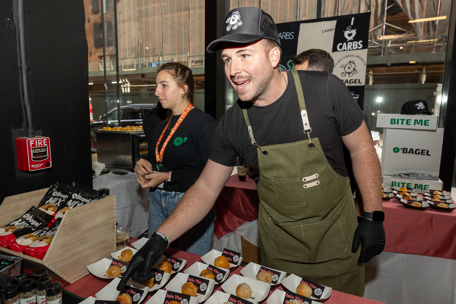 A man, from O’Bagel, in a black hat, shirt and green apron pointing at their parmesan and sun-dried tomato bagel bites filled with cream cheese.