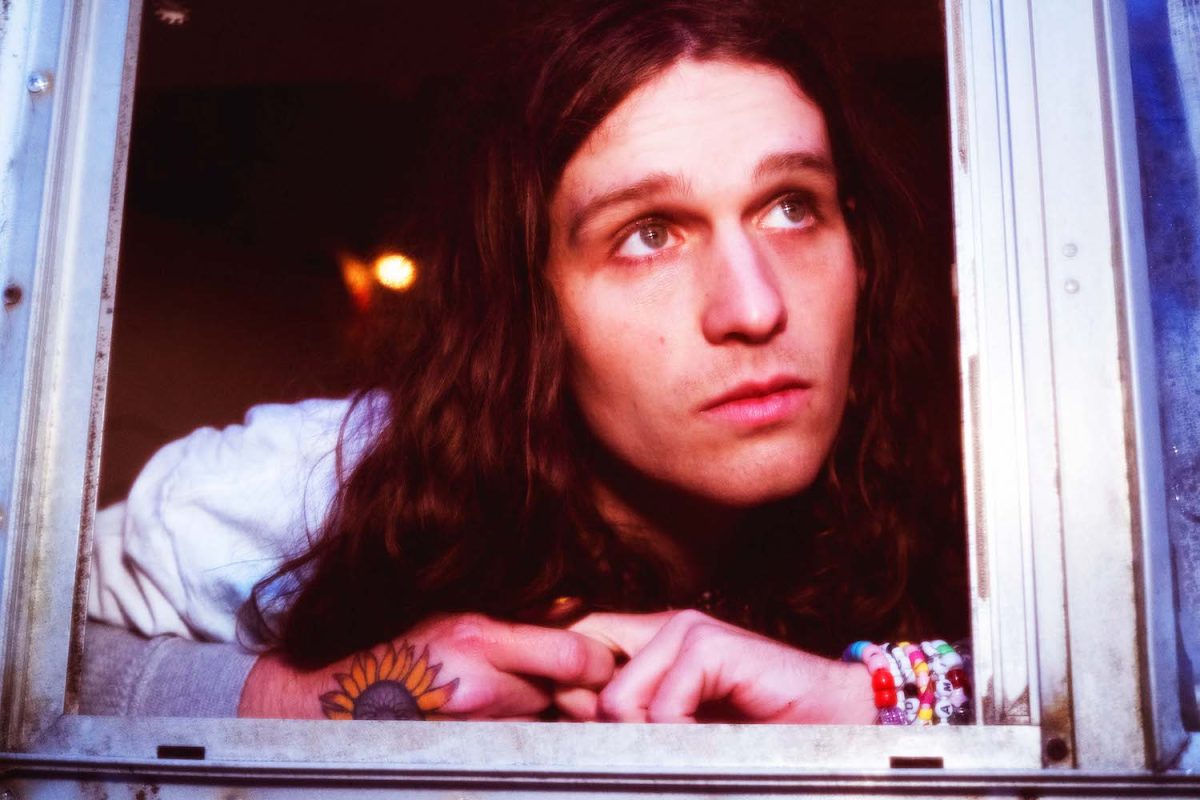 A person with long hair, a sunflower tattoo on his right hand, and a bead bracelet on his left hand looks out of the window.