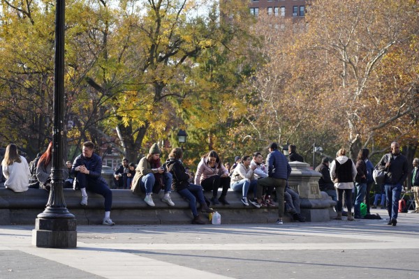 People sitting by the Washington Square fountain, with trees with green and yellow leaves behind them.