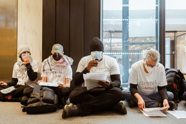 Four members of Faculty for Justice in Palestine, all wearing white shirts with the text “Ceasefire Now” written on them, sit on the second floor of the Bobst Library, reading statements into a remote microphone.