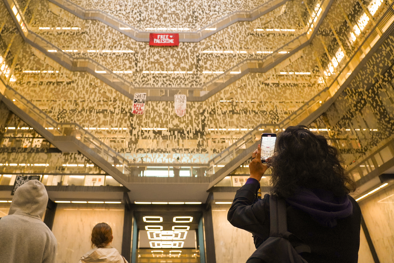 A woman with black hair wearing a black jacket takes photos of banners hanging from the staircases of the Bobst Library.