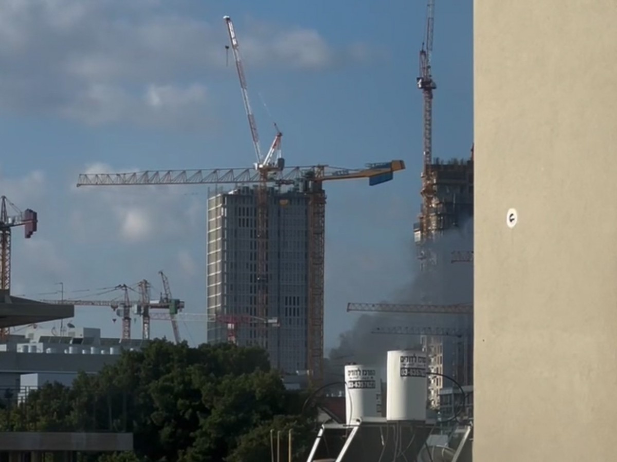 A view out of a window of a N.Y.U Tel Aviv residence hall showing smoke rising from the ground, among two buildings in construction and several cranes.