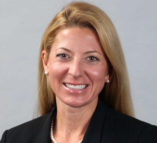 A woman with blonde hair is smiling. She is wearing pearl earrings and a pearl necklace.