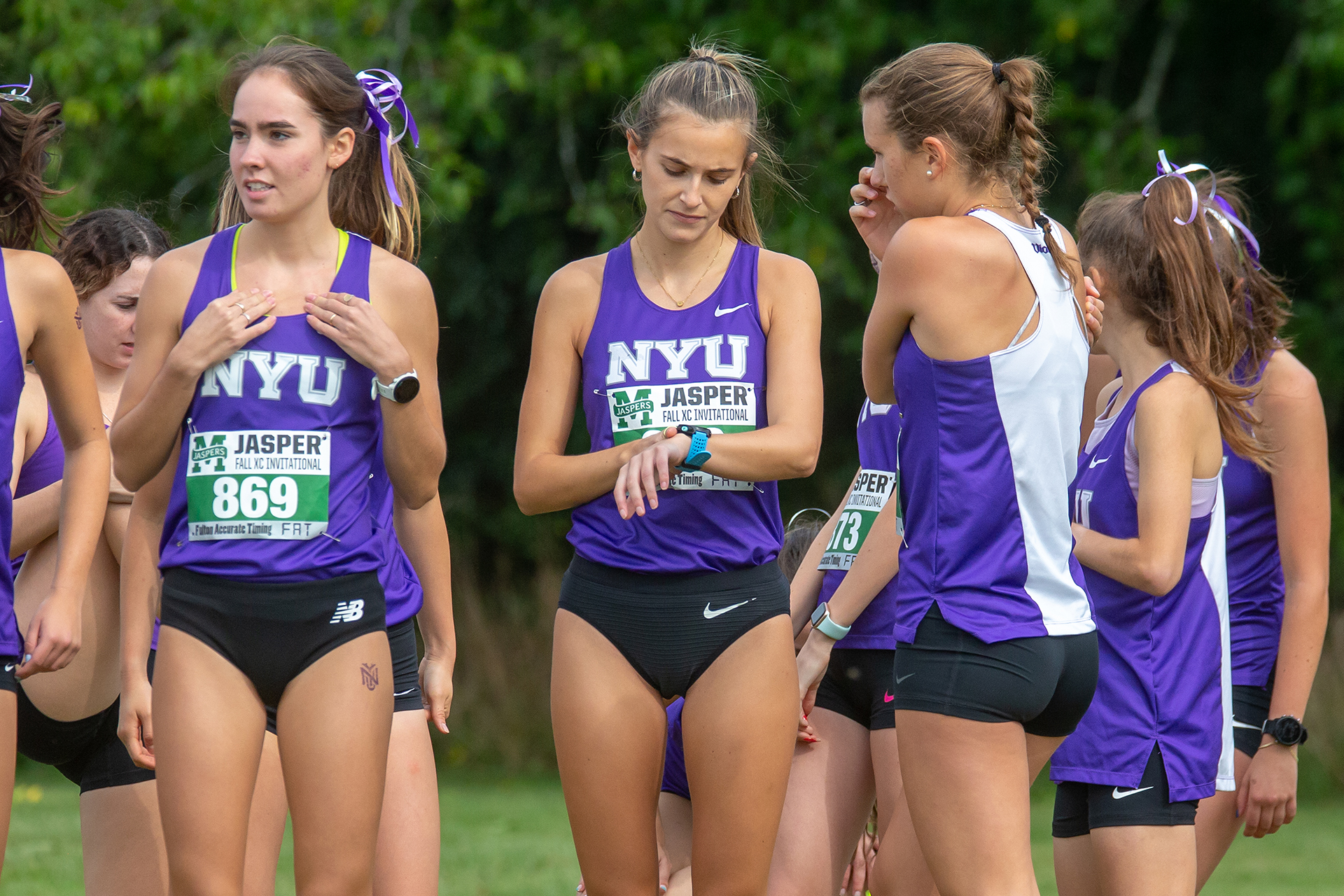 Multiple women wearing purple pinnies and black shorts standing in front of trees in the distance.