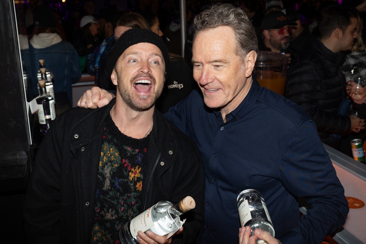 Aaron Paul in a black shirt and beanie and Bryan Cranston in a blue shirt, both holding bottles, smile with an arm around each other.