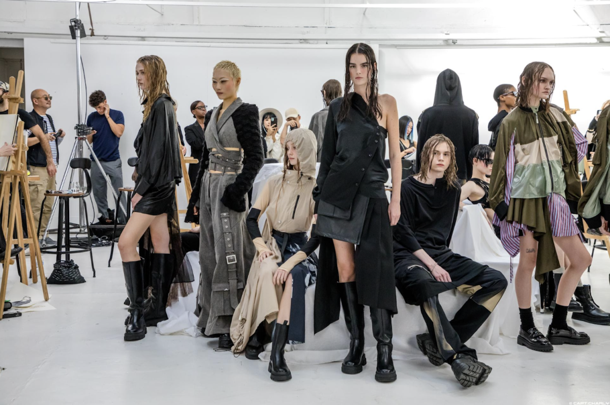Multiple models dressed in a variety of clothing are sitting, standing or posing. The background is white and to the left of the photo is a camera and a canvas.