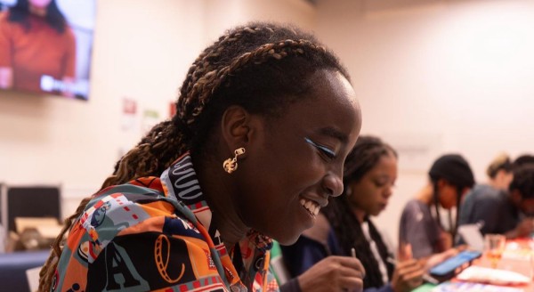 Someone with blue eyeliner, a gold earring, and a patterned shirt is looking down and smiling.