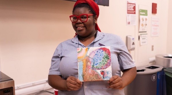 Someone wearing red glasses and a blue and white striped shirt is smiling and holding a painting.