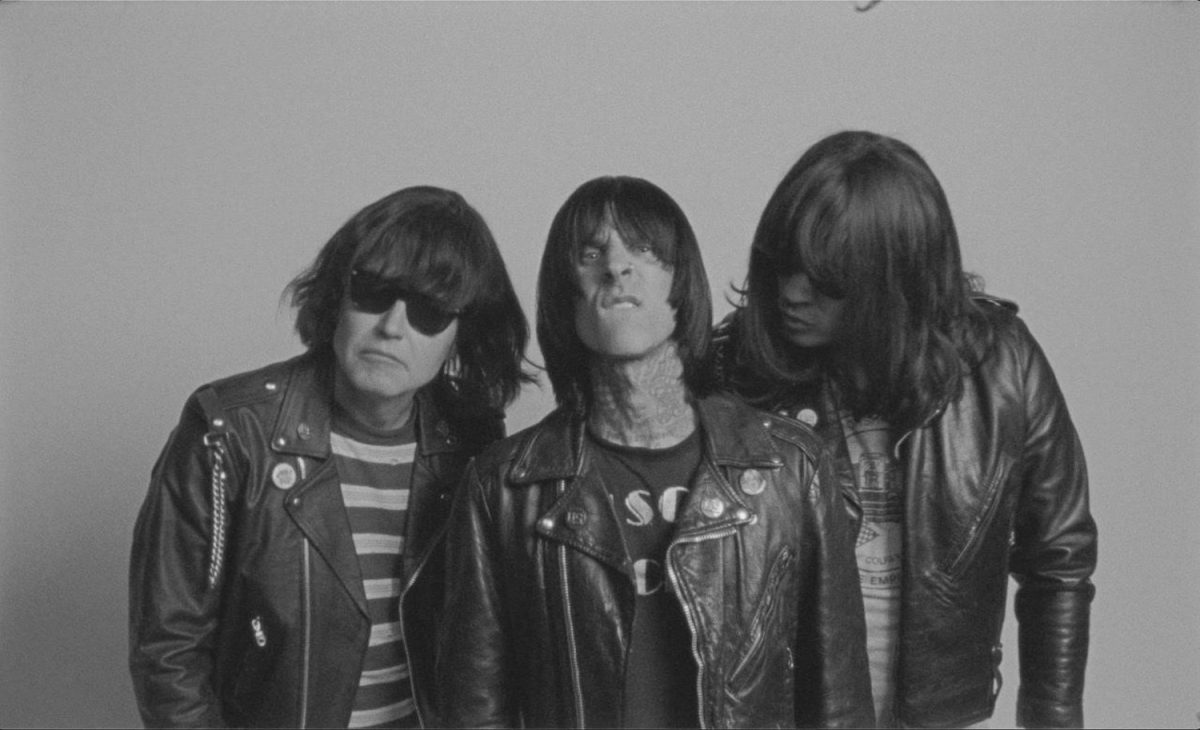 Three men wearing black leather jackets and with identical shaggy haircuts. Two are wearing glasses and one is facing the camera and showing his teeth.