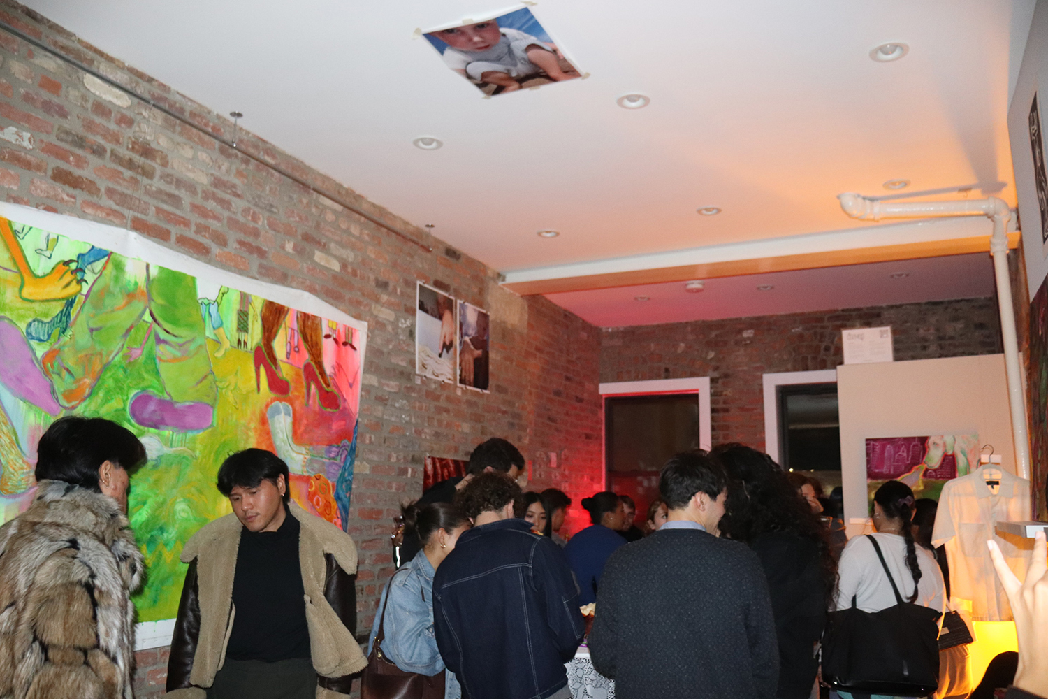 A crowd of people inside a room with brick walls on the left side and white walls on the other. A large painting of feet and legs in multiple colors is on the left side wall and there is a picture of a child on the white ceiling.