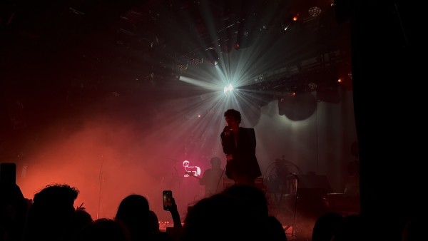 Someone is on stage, singing. There is a bright disco ball behind them and the silhouette of someone playing guitar. There is a red light and a red neon sign. There are people watching and one person is filming the singer with a phone.
