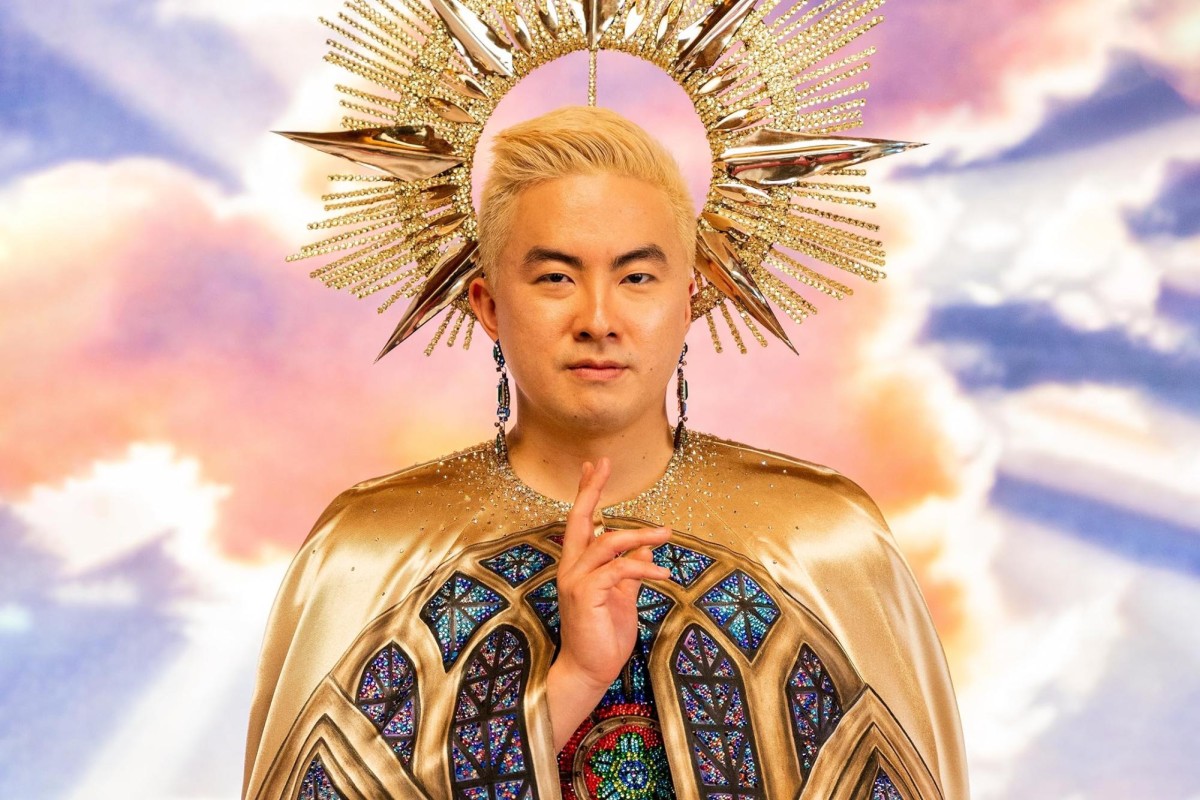 There is a person wearing a golden cloak with mosaic-stained glass art on it. They are raising two of their fingers. There is a round, gold headpiece behind their head and there are pink clouds in the background.