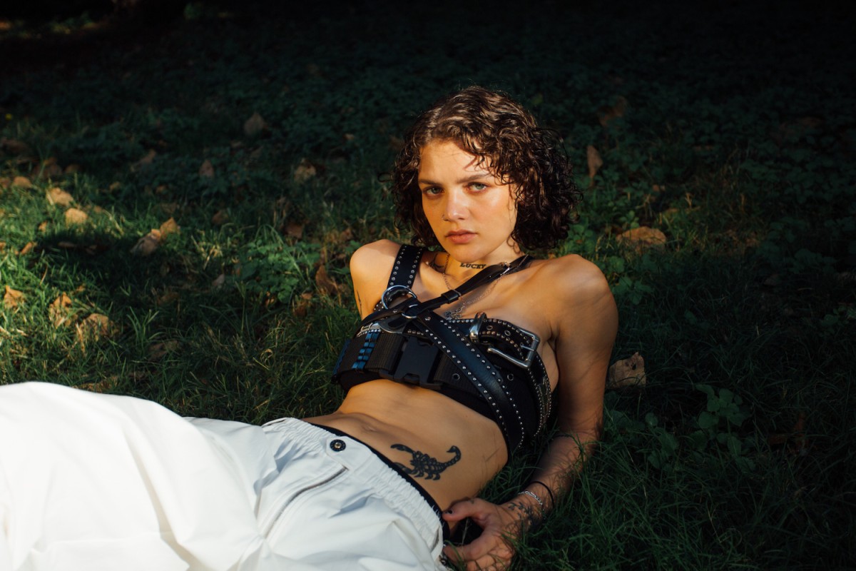 Someone with brown hair and blue eyes is lying down on grass. She is wearing white pants and a black, harness-like crop top.