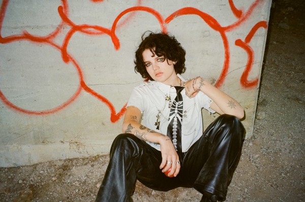 Someone with brown hair and blue eyes is sitting on the floor, leaning against a wall with red graffiti. She is wearing a white t-shirt with chains and a black tie with a white skeleton design on it. She is wearing black leather pants and has tattoos on her arms.